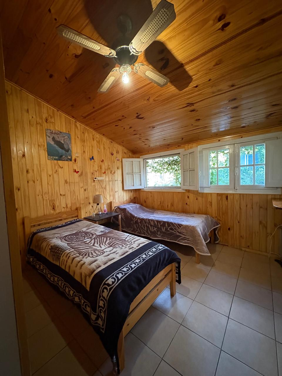 Segundo dormitorio con dos camas separadas, paredes revestidas en madera, ventanas y ventilador de techo.